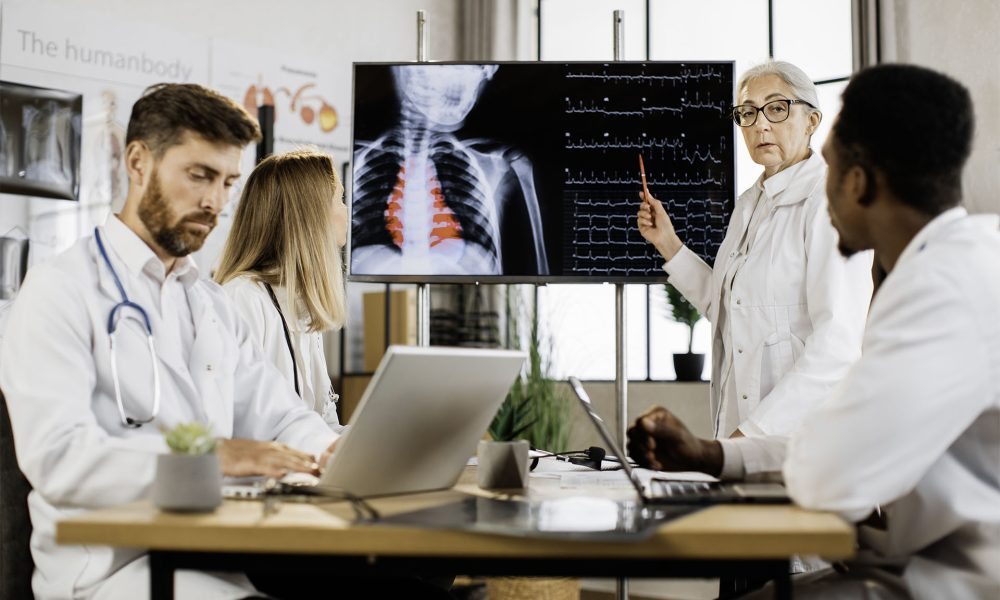 Mujer caucásica anciana con bata blanca de laboratorio de pie cerca de un gran monitor y hablando sobre las tasas de latidos del corazón durante la sesión informativa con médicos internacionales competentes. Concepto de medicina y atención médica.